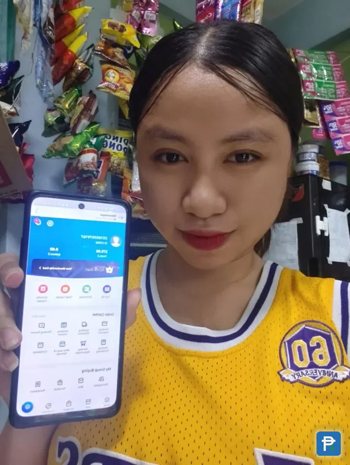 A shopper in a Philippine sari-sari store holding up a phone showing the Novaluxia PH app, with snack shelves behind her