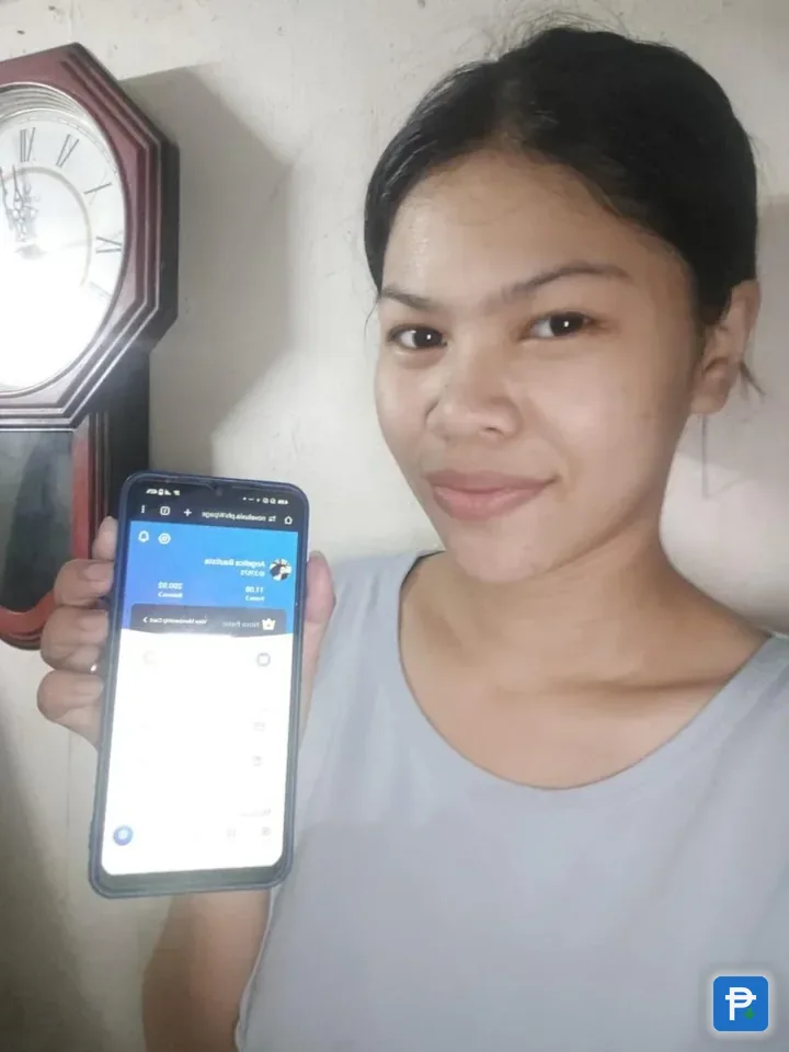 A user holding up a phone showing her Novaluxia PH wallet, with a wall clock behind her