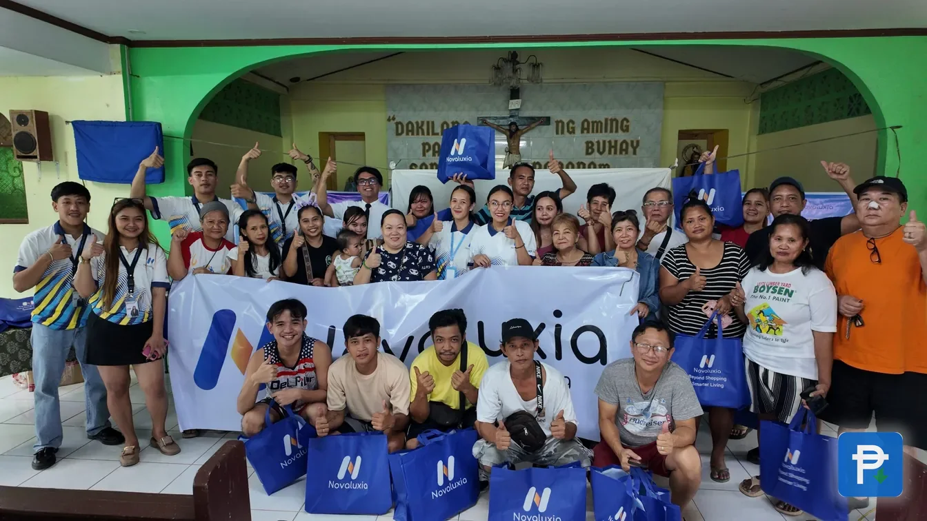 About forty Novaluxia PH community members in a hall, posing behind a branded banner with blue tote bags