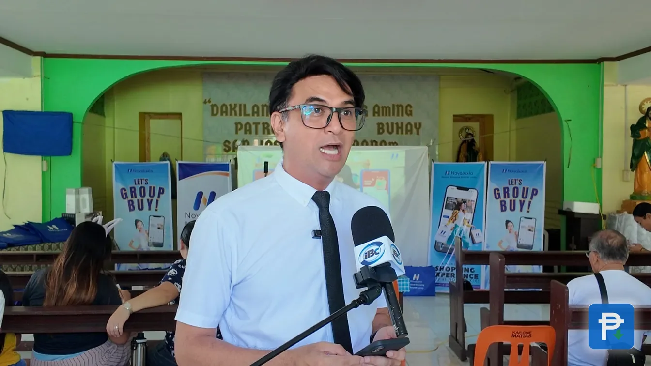 A speaker at the microphone during a Novaluxia PH group-buy orientation, posters on the wall behind him