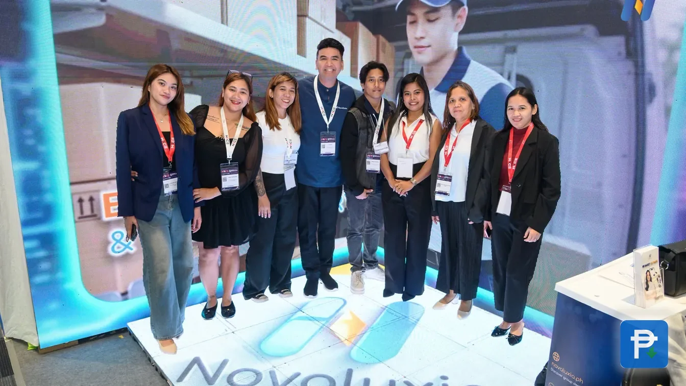 Eight Novaluxia staff in lanyards posing on a branded floor graphic at the expo booth