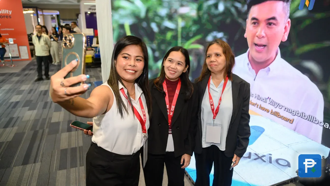 Three visitors taking a selfie in front of a Novaluxia expo booth screen, a dated public event photo for a legit-check file